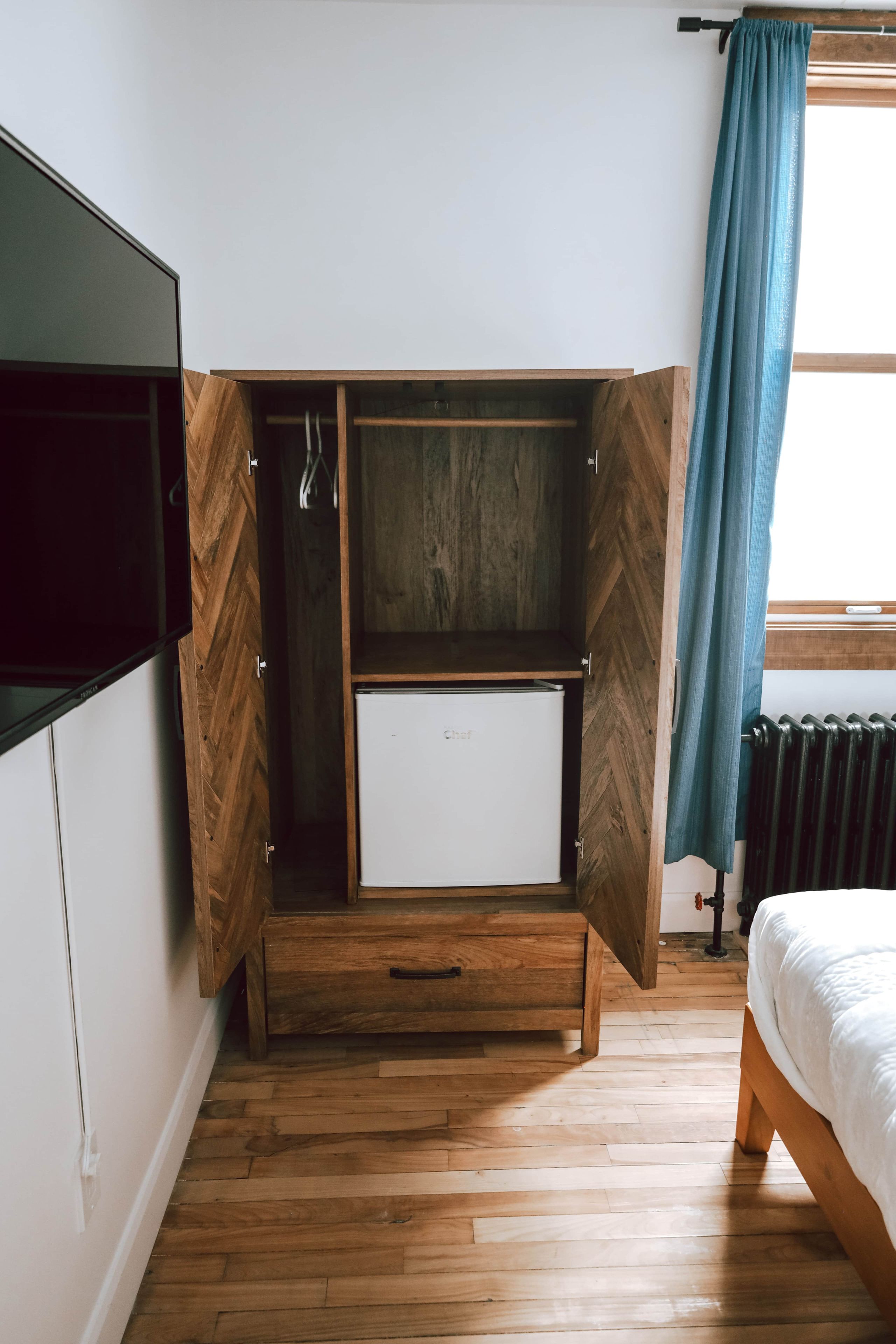 Una camera da letto con un letto, un armadio con mini-frigo integrato, pavimento in legno e una finestra con tende.