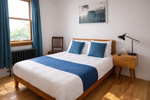 Ein Schlafzimmer mit Bett, Nachttisch, Lampe, Stuhl und einem Fenster mit Vorhängen.