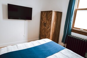 Un dormitorio con cama, armario, televisor montado en la pared y ventana con cortinas.