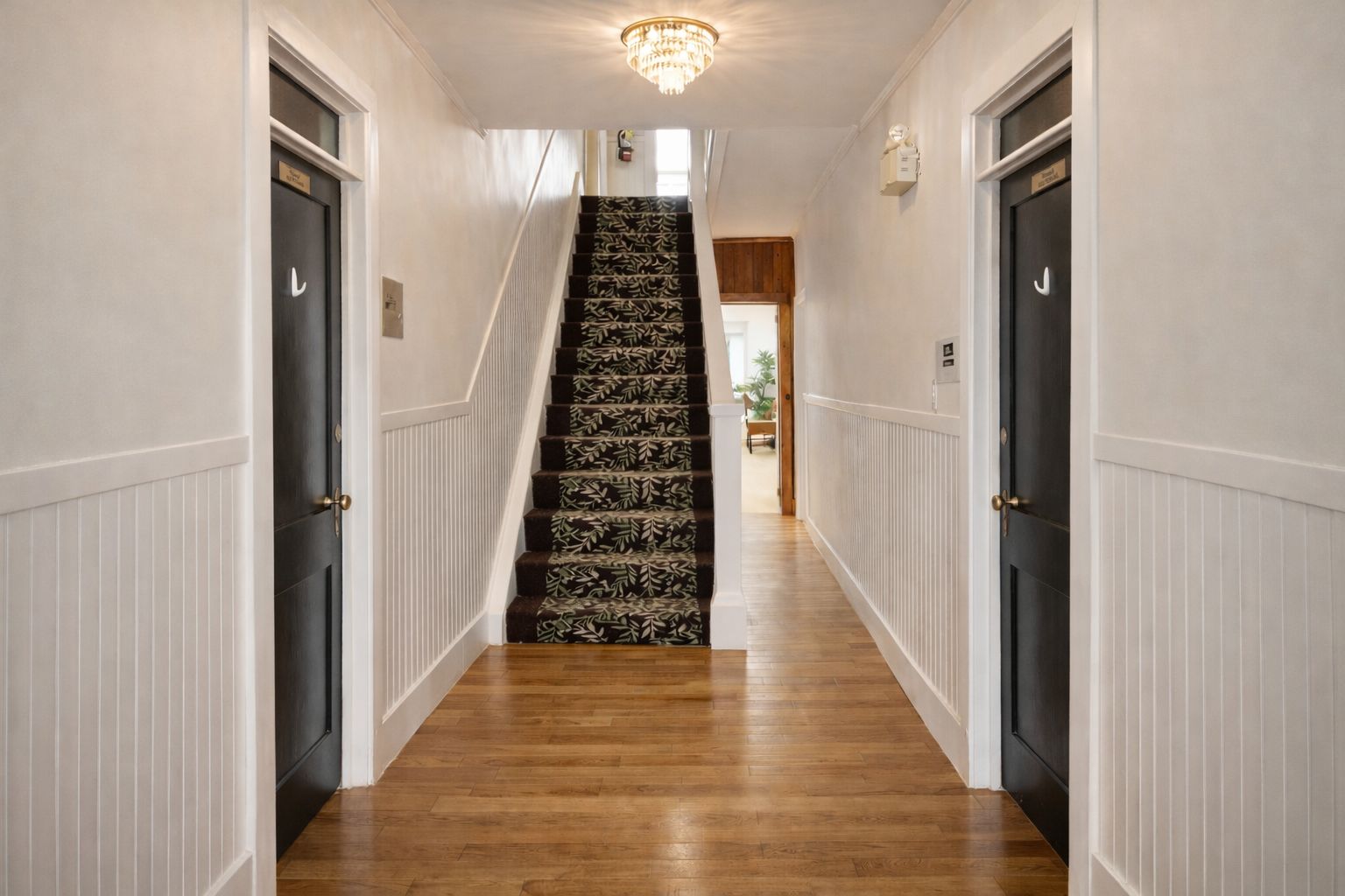 A hallway with a wooden floor, white wainscoting, and a staircase with patterned carpet.
