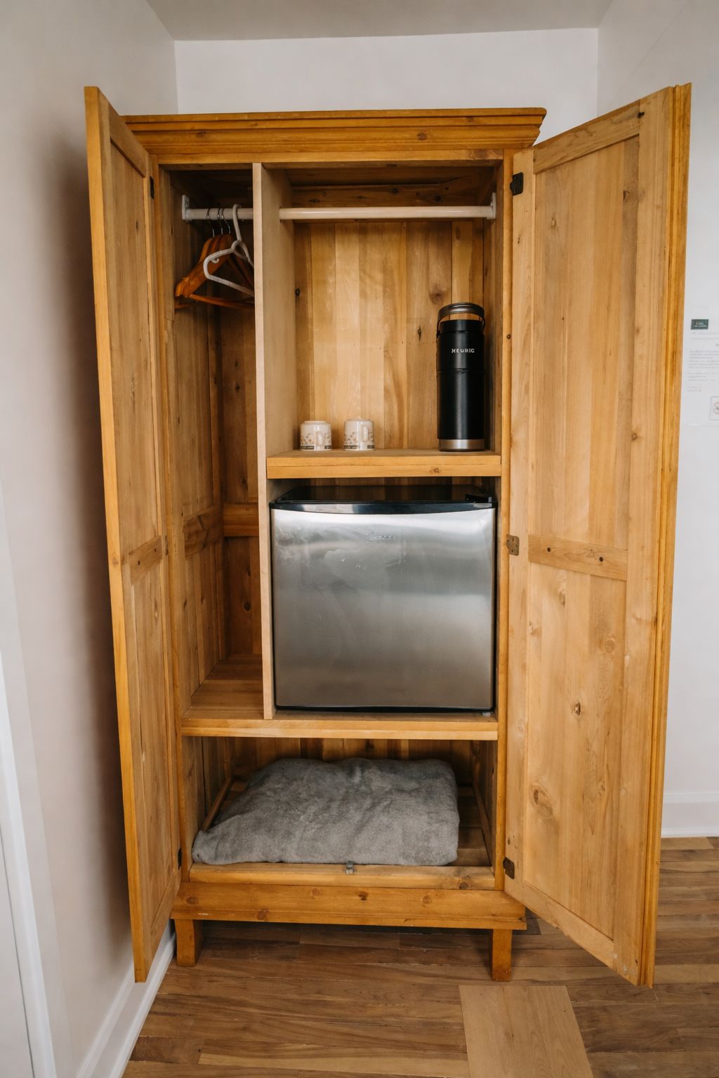 A wooden cabinet with a built-in refrigerator, a shelf with two cups, and a hanging rack.