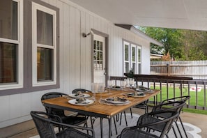 An outdoor dining area with a wooden table set for four, surrounded by black chairs, against a white exterior wall with windows.