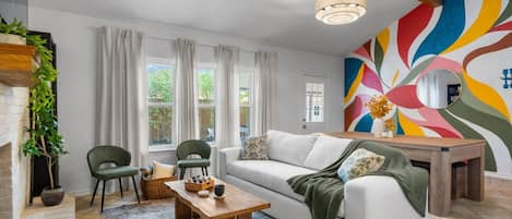 A modern living room with a white sofa, two green chairs, a wooden coffee table, and a colorful abstract wall mural.