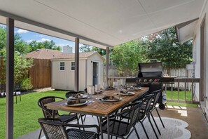Patio esterno con tavolo in legno apparecchiato per mangiare, circondato da sedie e una griglia barbecue sullo sfondo.
