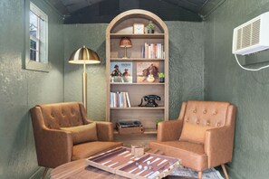 Ein Zimmer mit zwei Ledersesseln, einem Bücherregal, einer Lampe und einem Tisch mit einem Gesellschaftsspiel.