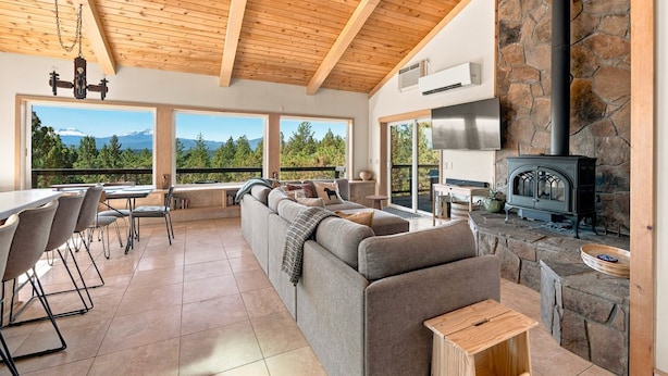 A spacious living area with a stone fireplace, large windows offering a mountain view, and a modern kitchen with a stainless steel range. - Cascade Views | Family Retreat | Dog Friendly (Sisters)