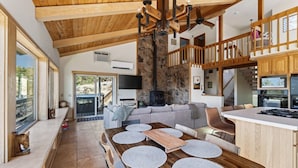 A spacious living area with a high ceiling, a stone fireplace, a wooden dining table with placemats, and a view through large windows. - Cascade Views | Family Retreat | Dog Friendly (Sisters)