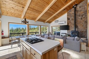 A modern kitchen with a central island, a stone fireplace, and a large window overlooking a forested area.