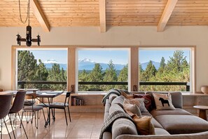 A modern living room with a sofa, chairs, a dining table, and large windows offering a mountain view.