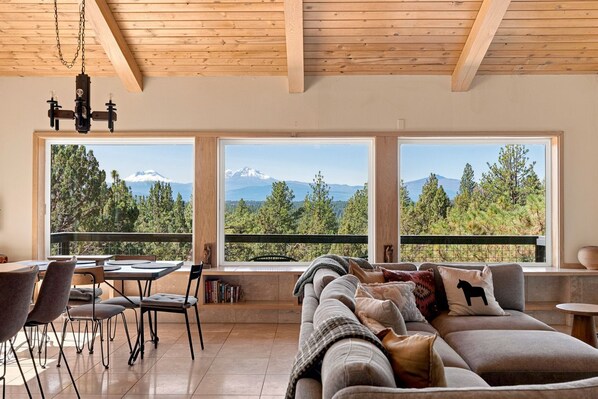 A modern living room with a sofa, chairs, a dining table, and large windows offering a mountain view.
