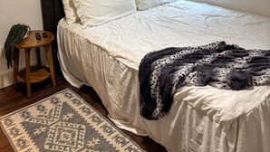 A bedroom with a bed, a bedside table, a window with blinds, and a patterned rug. - Downtown Bryan Apt - Walk to Everything (Bryan)