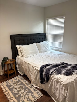 A bedroom with a bed, a bedside table, a window with blinds, and a patterned rug. - Downtown Bryan Apt - Walk to Everything (Bryan)