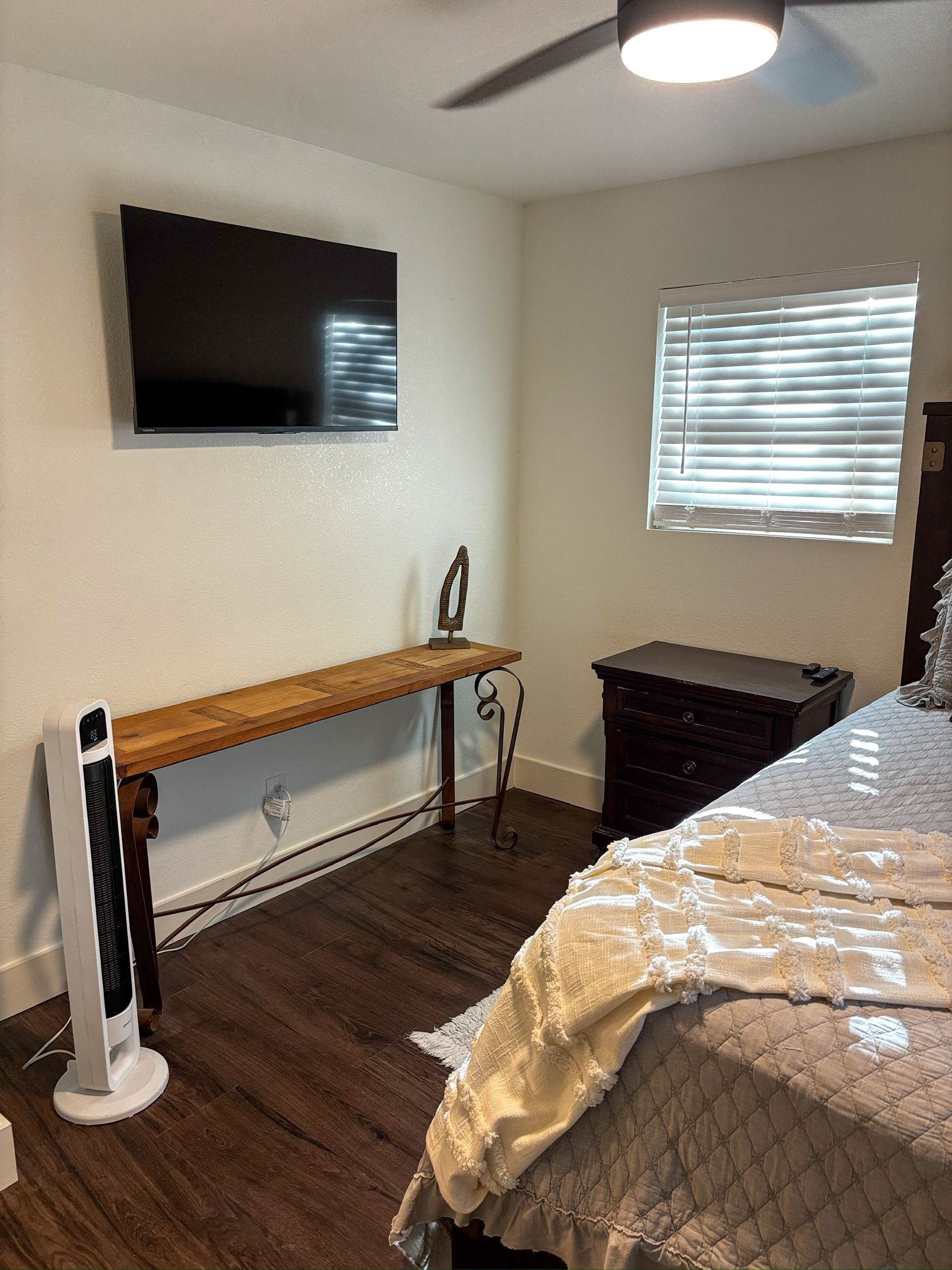 Un dormitorio con cama, televisor montado en la pared, ventana con persianas y un calefactor en el suelo.