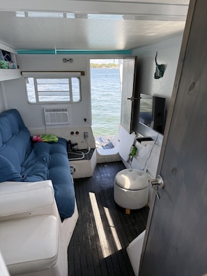 A boat interior with a blue cushioned seating area, a small table, a television, and a window overlooking the water.