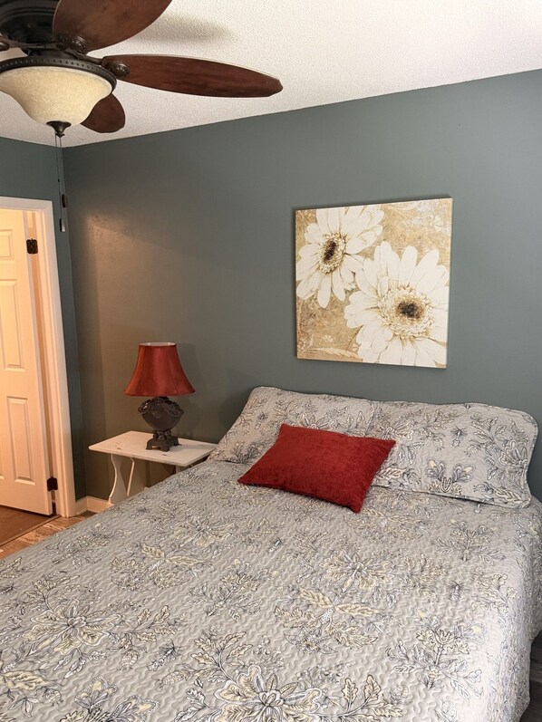 A bedroom with a bed, a nightstand, a lamp, a ceiling fan, and a wall-mounted artwork.
