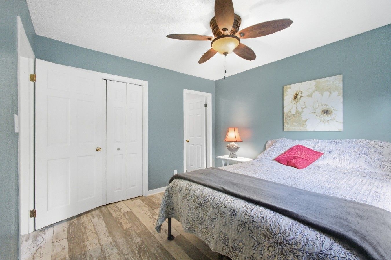 Une chambre à coucher avec un lit, un ventilateur de plafond, une table de chevet avec une lampe et un mur orné d’un tableau représentant des motifs floraux.