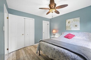 A bedroom with a bed, a ceiling fan, a nightstand with a lamp, and a wall with a floral painting.
