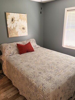 Un dormitorio con cama, almohada roja, un cuadro de flores en la pared y una ventana con persianas.