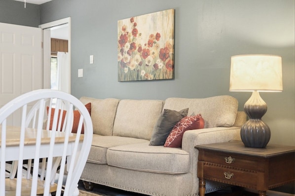 A living room with a beige sofa, a white chair, a wooden nightstand, and a table lamp.