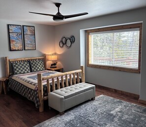 Ein Schlafzimmer mit einem hölzernen Bett, einer Bank, einem Deckenventilator, einem Fenster mit Jalousien und zwei Bildern an der Wand.