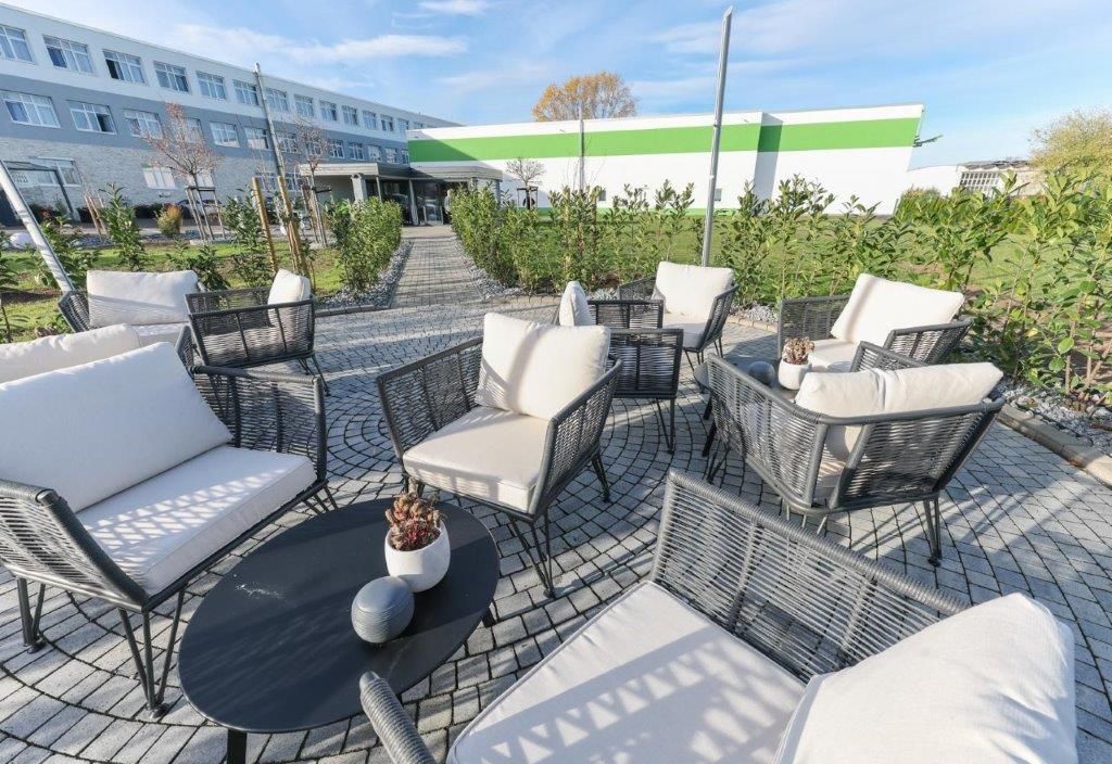 An outdoor seating area with wicker furniture and a round table, surrounded by greenery and a building in the background.