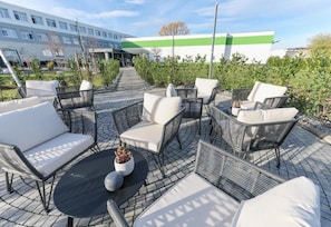 An outdoor seating area with wicker furniture and a round table, surrounded by greenery and a building in the background.
