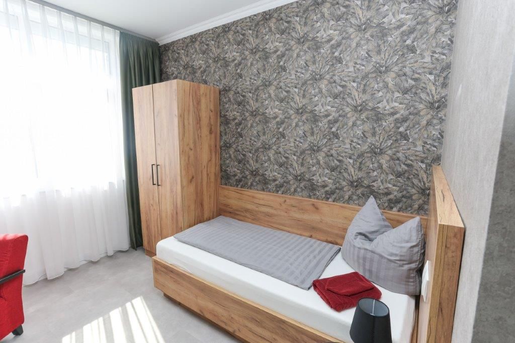 Quarto individual com cama de solteiro, cabeceira de madeira, colchão cinza e uma cadeira vermelha.