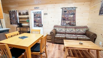 Interior de cabana com mesa de jantar de madeira, cadeiras, sofá e pequena mesa de madeira.