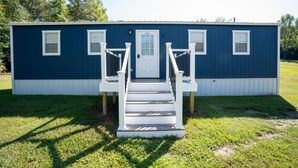 Een gelijkvloers blauw huis met een witte voordeur, twee ramen aan de zijkant en een klein portaaltje met treden naar de deur.