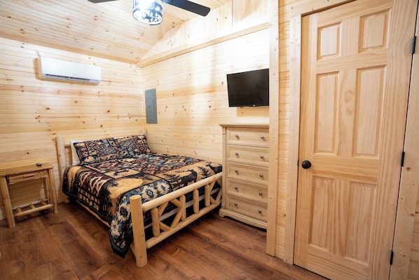 A wooden-paneled room with a bed, a dresser, a television, and a door. - Cozy 2-bedroom cabin with AC in Jacksonville, Alabama (Jacksonville)