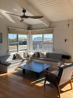 A sunlit room with a ceiling fan, large windows, a built-in bench with cushions, a coffee table, and a chair. - Renovated 1800 sq ft house, AC throughout, Fios, Large Deck (Davis park)