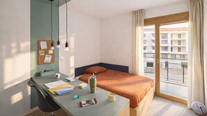 Une chambre d’hôtel comprenant un lit, un bureau avec une chaise, un tableau d’affichage et une fenêtre donnant sur des immeubles.