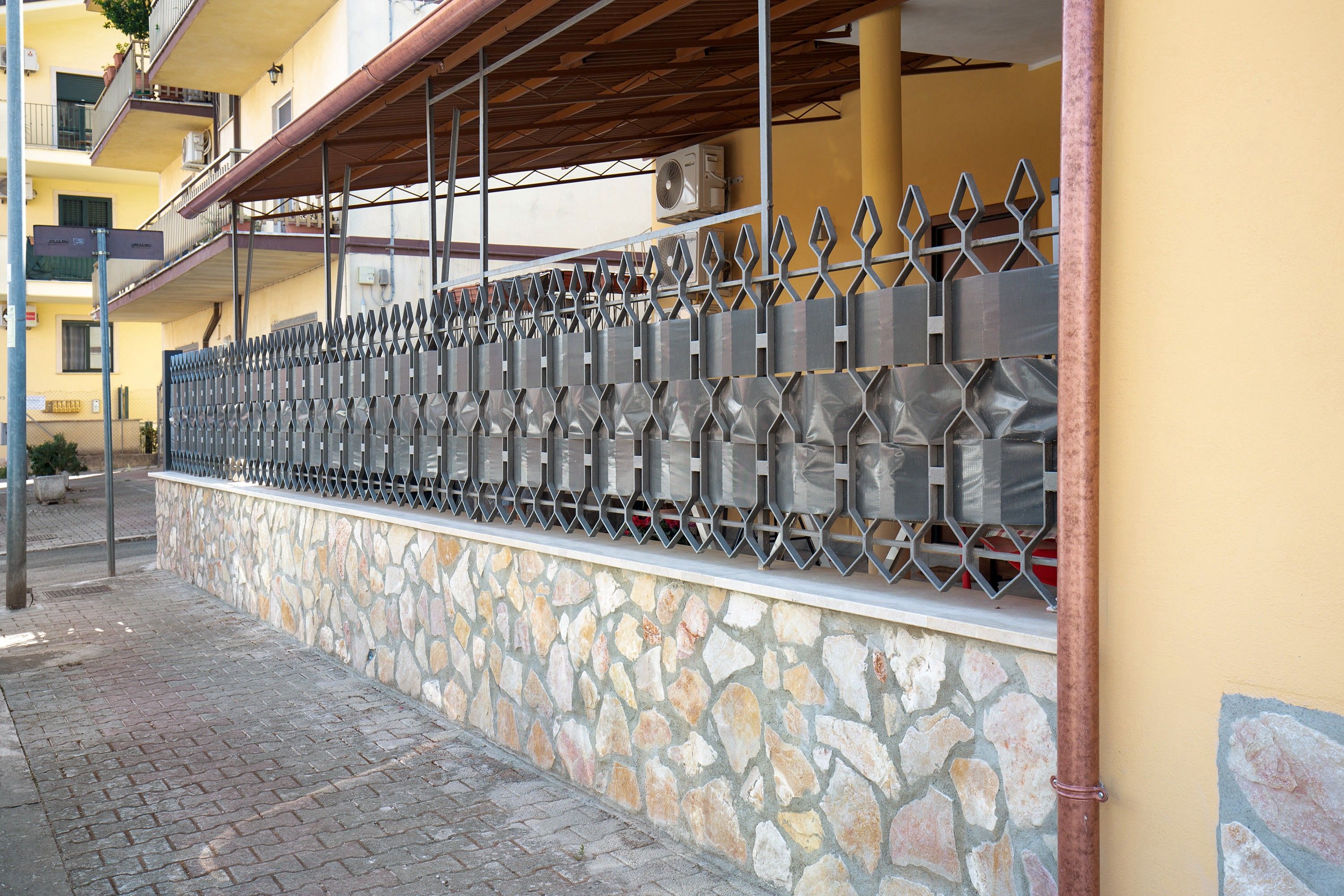 A stone wall with a metal fence, a paved sidewalk, and a yellow building with balconies.