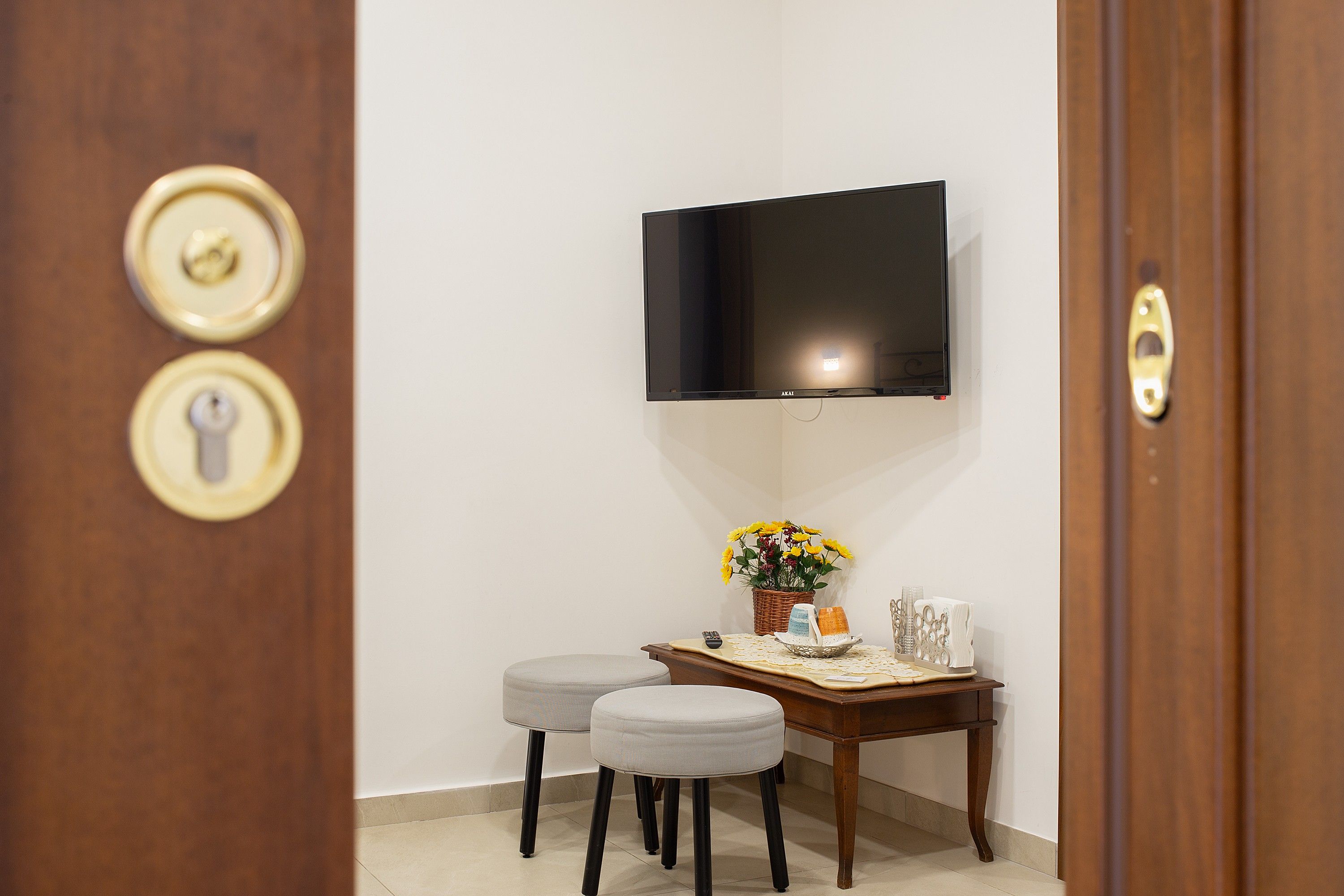 A room with a flat-screen TV mounted on the wall, a small wooden table with a vase of flowers, two grey ottomans, and a door with a keyhole.