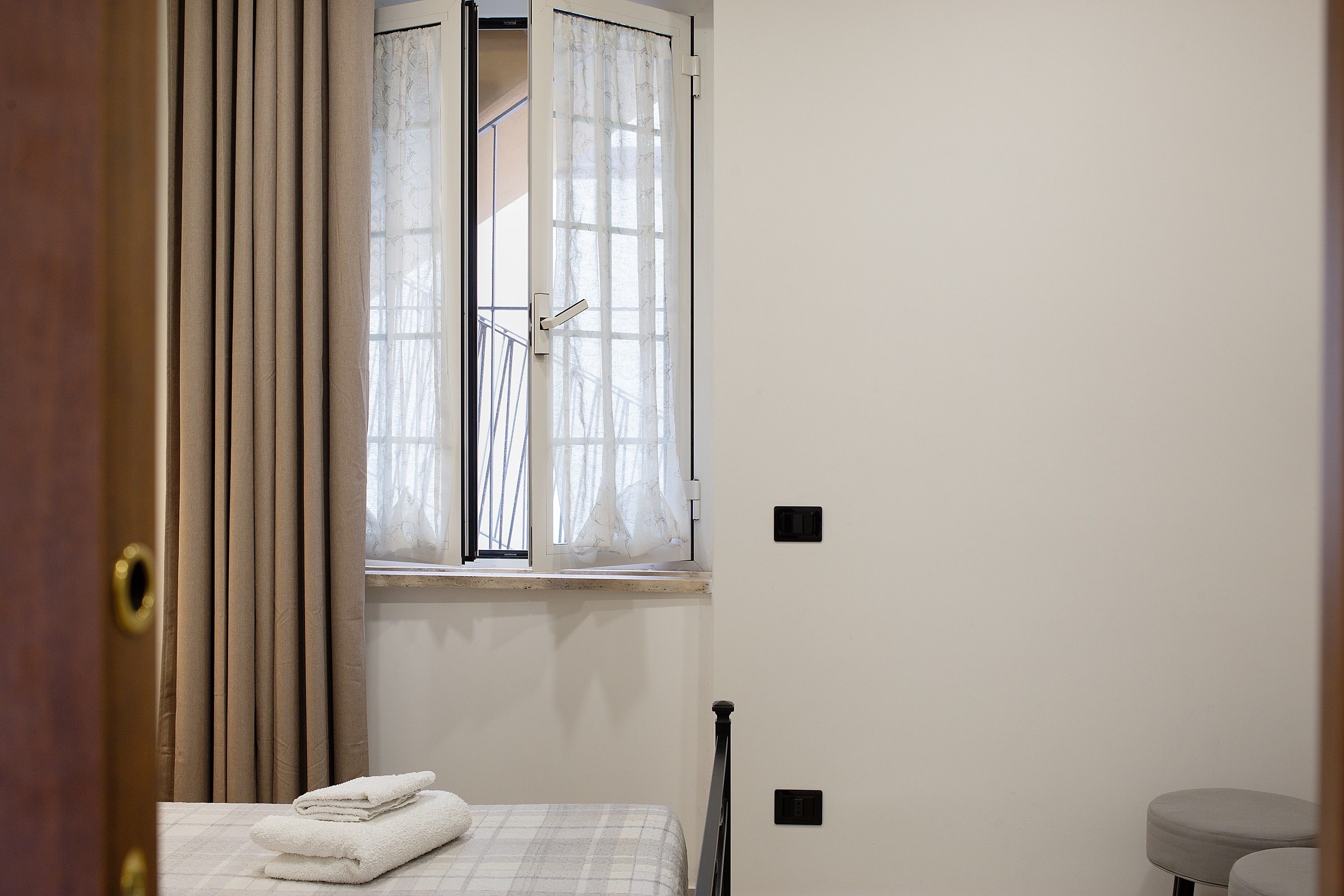 Una habitación con una cama, una ventana con cortinas, una puerta y un taburete pequeño.