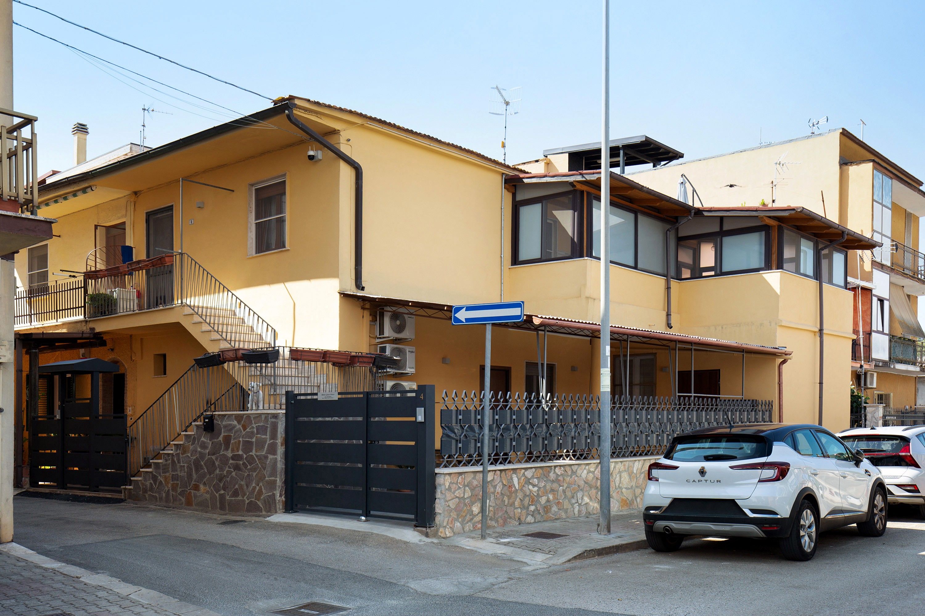 A yellow building with a black gate, a staircase, and a parked white car.