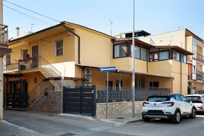 A yellow building with a black gate, a staircase, and a parked white car.