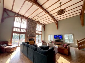 A spacious living room with a stone fireplace, large windows, and a television mounted on the wall.