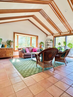A spacious living room with a high ceiling, large windows, and a variety of seating options including a sofa and wicker chairs.