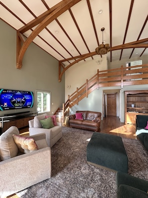 A spacious living room with a high ceiling, wooden beams, a staircase, and a television displaying a game. - Pen y Bryn - 5BR Blue Ridge Luxury Retreat with Pool, Library & Game Room (Faber)