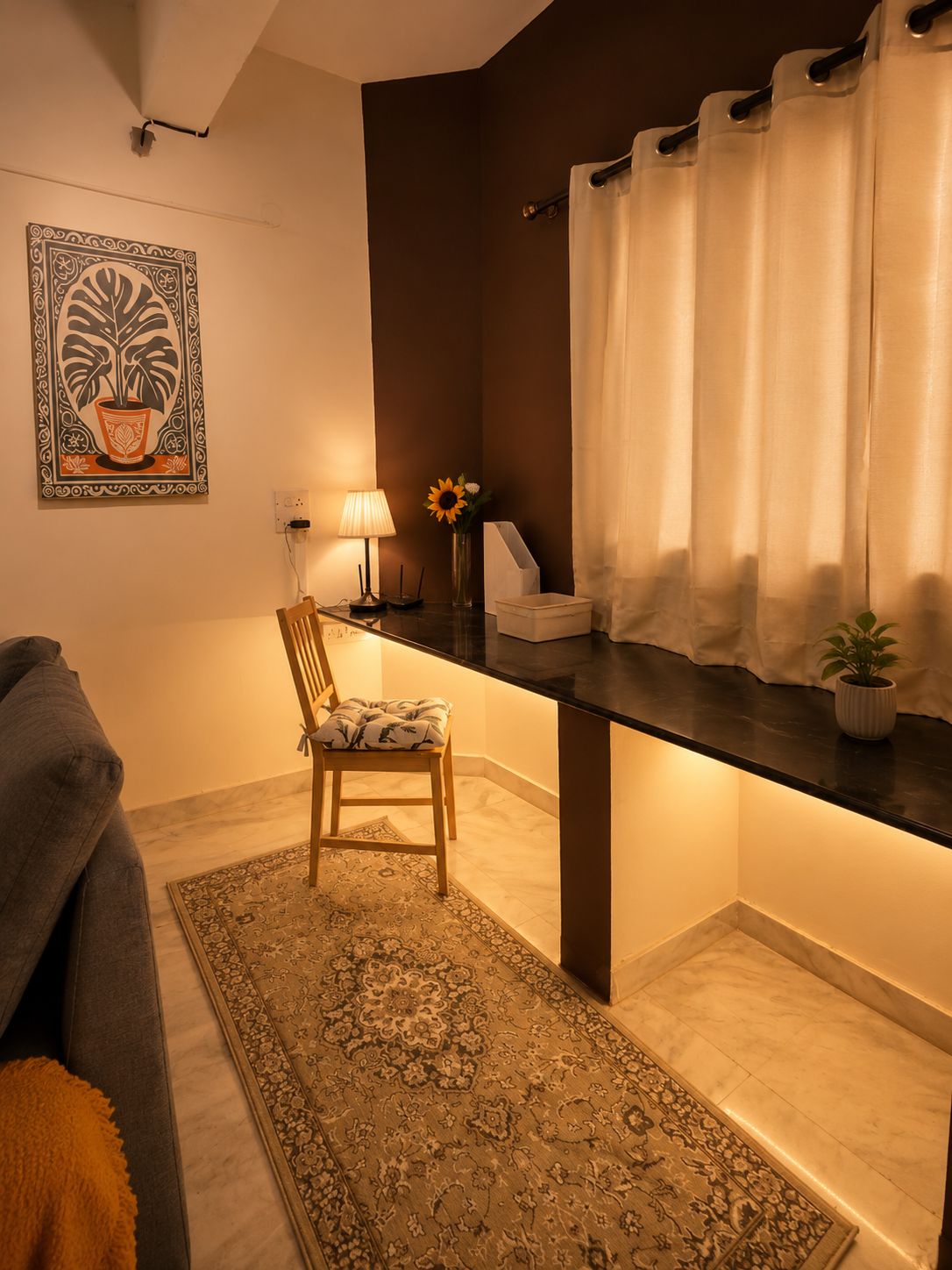 Una habitación con una pared de acento marrón oscuro, una cortina blanca, una silla de madera, una mesita con una lámpara, un jarrón con flores y una alfombra estampada.