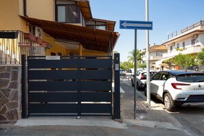 Una entrada protegida por un muro de piedra, con un cartel azul de indicaciones y varios coches aparcados.