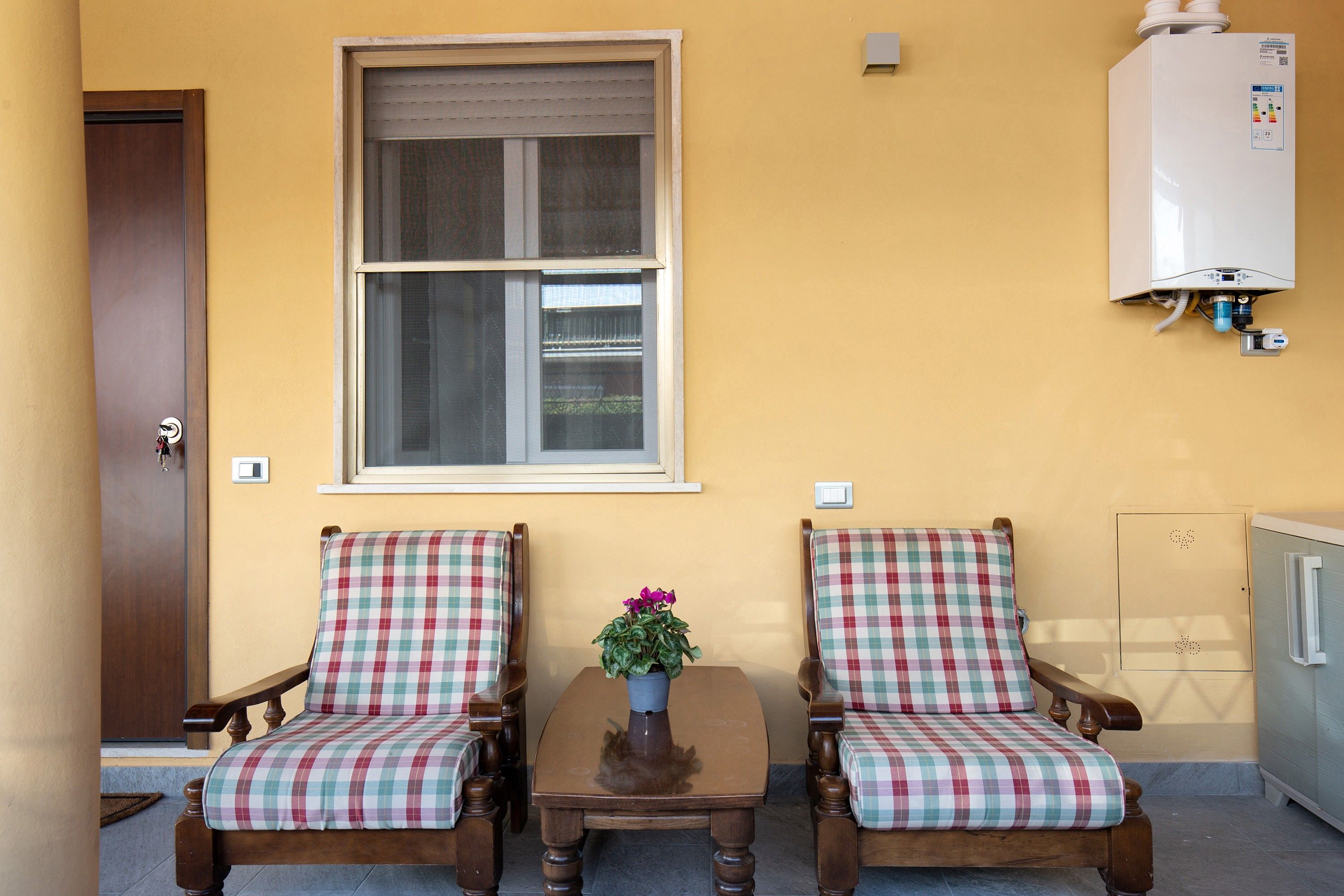 Una habitación con dos sillas de madera con cojines escoceses, una mesita con un florero y una ventana con vista a un edificio.