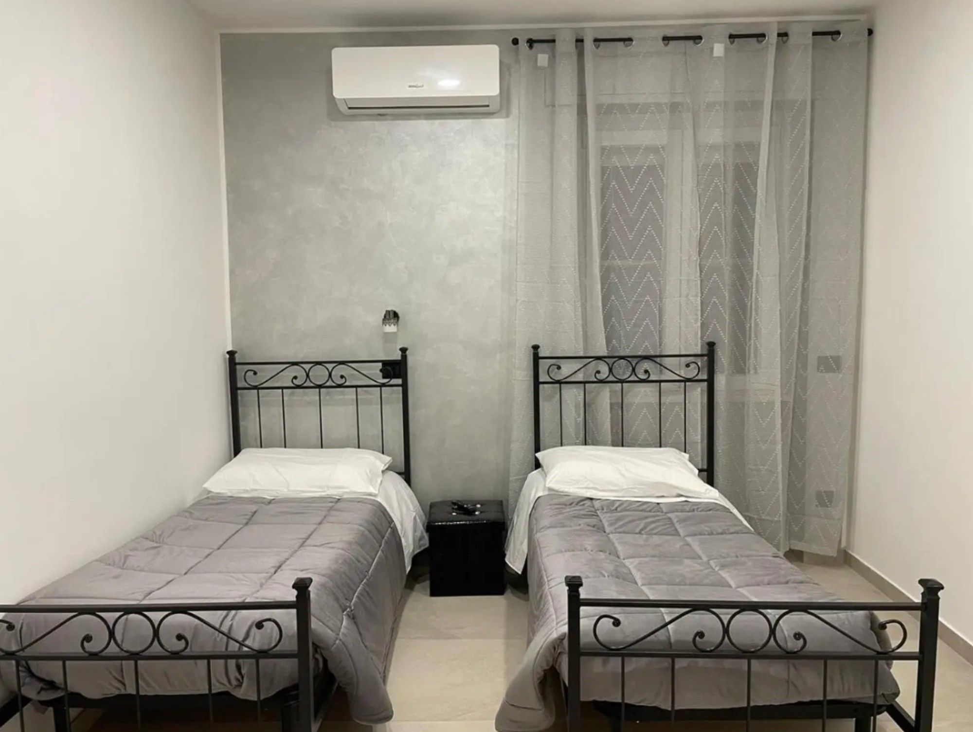 Ein Zimmer mit zwei Einzelbetten, einem kleinen Nachttisch, einer Wandklimaanlage und einem Fenster mit transparenten Vorhängen.