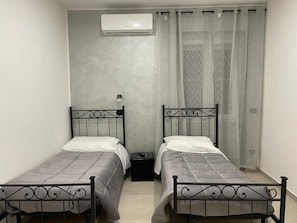 Une chambre avec deux lits simples, une petite table de chevet, un climatiseur fixé au mur et une fenêtre avec des rideaux légers.