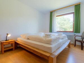 A bedroom with a wooden bed, a bedside table with a lamp, a chair, and a window with curtains. - Holiday Apartment With 2 Bedrooms (St. Peter-Ording)