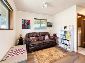 Ein Wohnzimmer mit einem braunen Ledersofa, einem Schachbrett, einem Bücherregal und einem Fenster mit Blick auf Grünflächen.