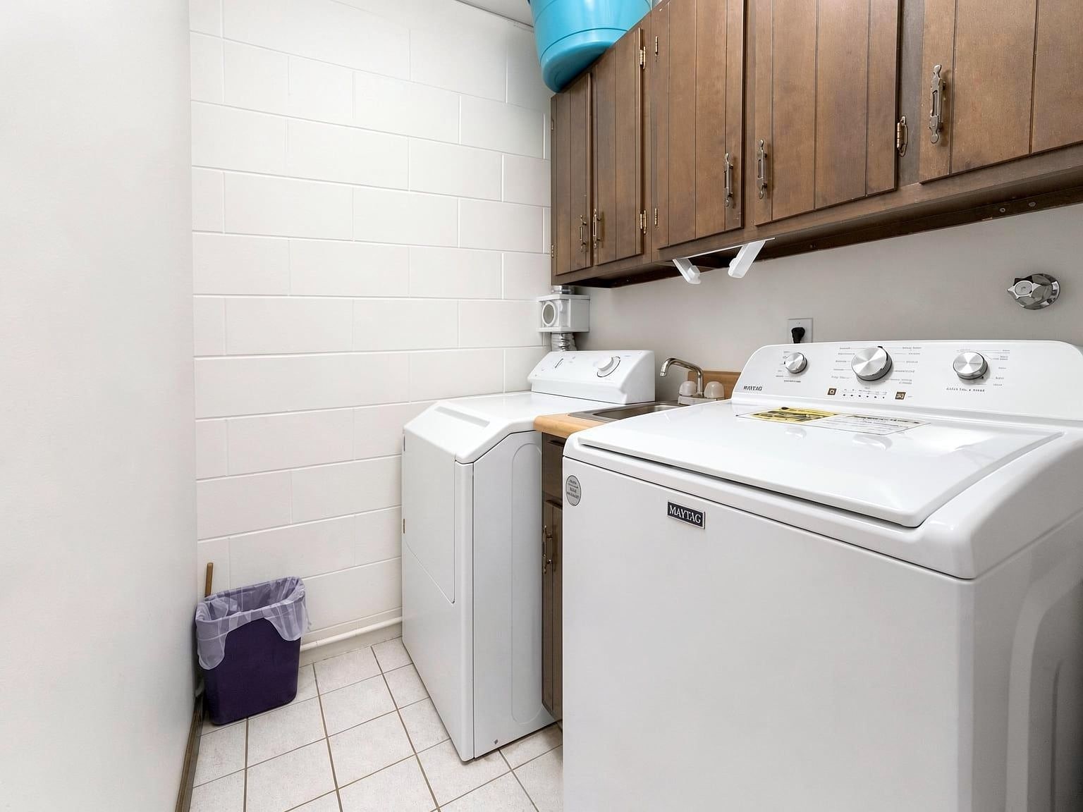A compact laundry room with a front-loading washer and dryer, wooden cabinets above, and a blue water tank on the ceiling.