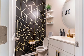 Ein modernes Badezimmer mit Wand in geometrischem Muster, Toilette, Waschbecken mit goldfarbenen Armaturen und einer kleinen Pflanze auf einem Regal.