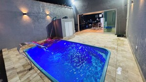 A swimming pool with a tiled deck, a covered patio area, and a view into a kitchen. - Perfect com piscina e 3-bedroom house in Peruíbe for a relaxing getaway (Peruíbe)